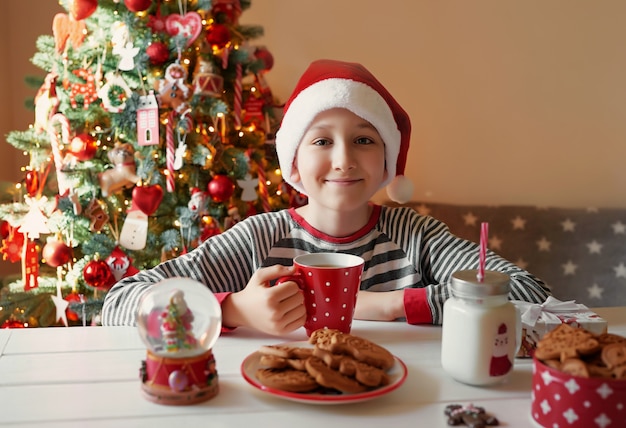 クリスマスツリーでお茶のクリスマス赤カップを持つ少年の笑顔。子供連れの家族は冬休みを祝います。自宅でクリスマスイブ。クリスマスキッチンの子少年。サンタ帽子の少年。