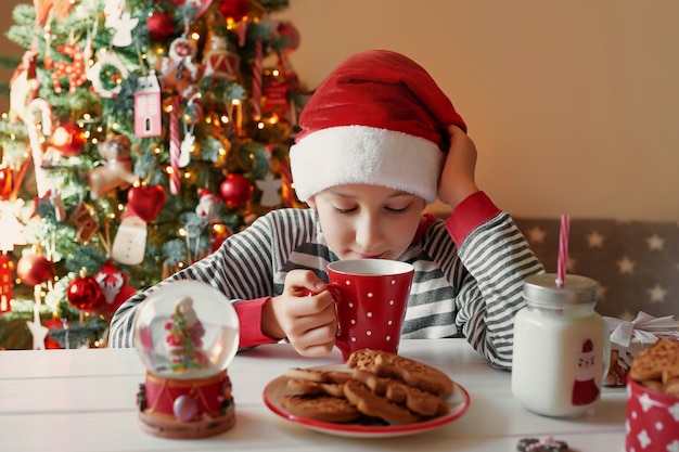 Smiling boy with christmas red cup of tea at christmass tree . Family with kids celebrate winter holidays. Christmas eve at home. Child boy in christmas kitchen. Boy in Santa hat.