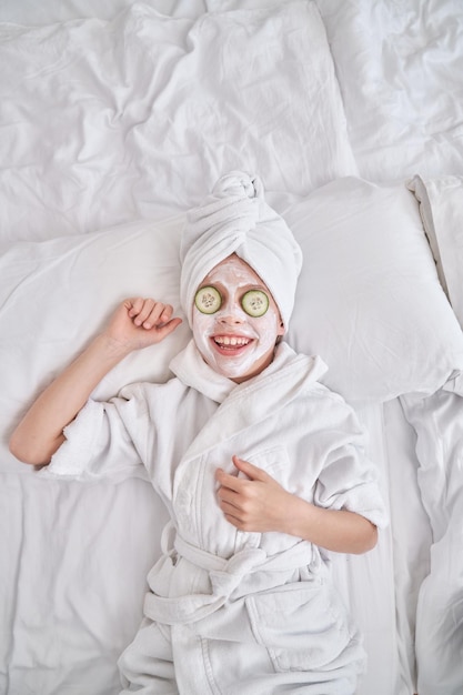 Smiling boy with facial mask and cucumber slices on eyes wearing white towel turban while lying on bed during spa procedures in bedroom at home