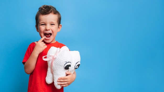 A smiling boy with healthy teeth holds a plush tooth in his\
hands on a blue isolated background oral hygiene pediatric\
dentistry prevention of caries a place for your text