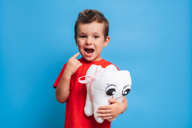 A smiling boy with healthy teeth holds a plush tooth in his hands on a blue isolated background oral hygiene pediatric dentistry prevention of caries