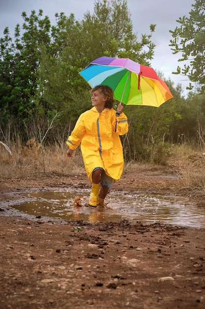 Smiling boy with rainbow umbrella playing in puddle