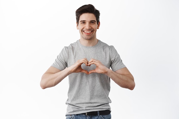 Smiling boyfriend shows heart sign, I love you gesture, express sympathy and like, look heartfelt, stands against white background