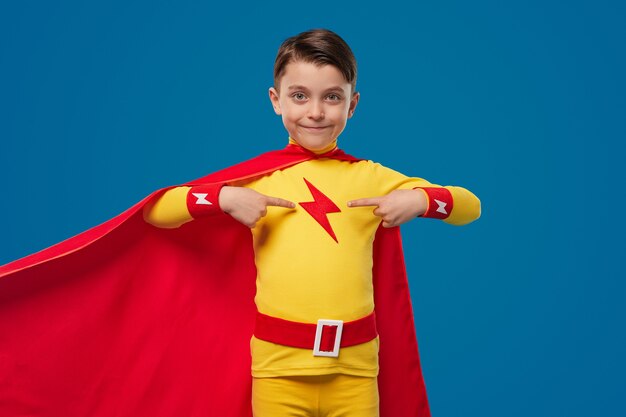 Smiling brave boy in superhero costume with red cape pointing at himself as being proud and ready to help while standing against blue background