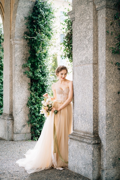 Smiling bride in a dress with a bouquet of flowers stands near the arch of an ancient villa lake