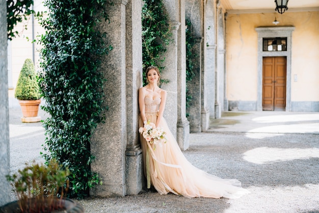 Smiling bride in a dress with a bouquet of pink flowers leaning against a pillar in a vaulted room