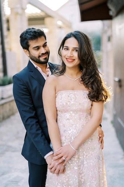 Photo smiling bride holds the hand of groom looking at her hugging her from behind near an old house