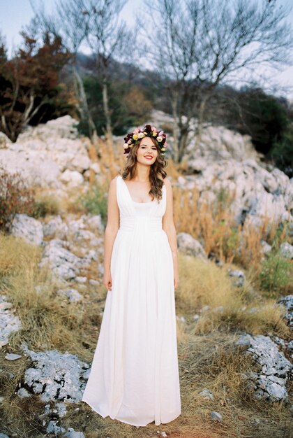 Smiling bride in a multicolored wreath stands on a mountain among the stones