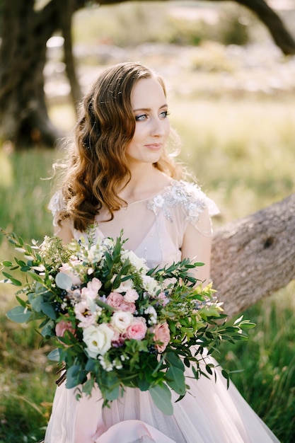 Smiling bride sits on a tree trunk with a bouquet of flowers