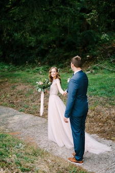 La sposa sorridente con un bouquet tira la mano dello sposo lungo il sentiero in giardino