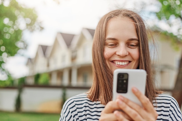 Smiling brown haired woman browsing internet on smart phone standing outdoor in street with houses on background checking social network while walking outside