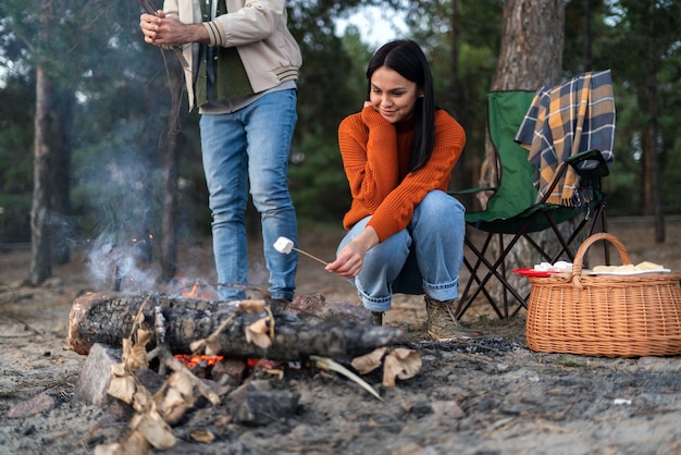 Smiling brunette caucasian woman camping and holding marshmallows at the stick while sitting at the nature with her friend or husband at the background