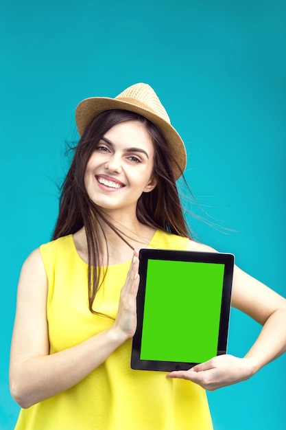 Smiling brunette girl dressed in yellow shirt and hat holds tablet computer with green screen before blue background advertise space concept