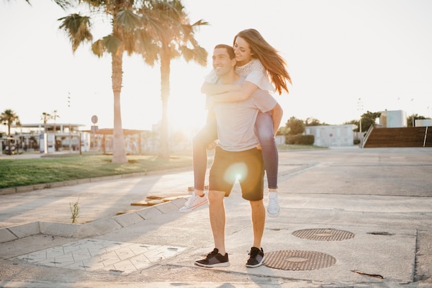 A smiling brunette girl is riding her Hispanic boyfriend between palms in Spain