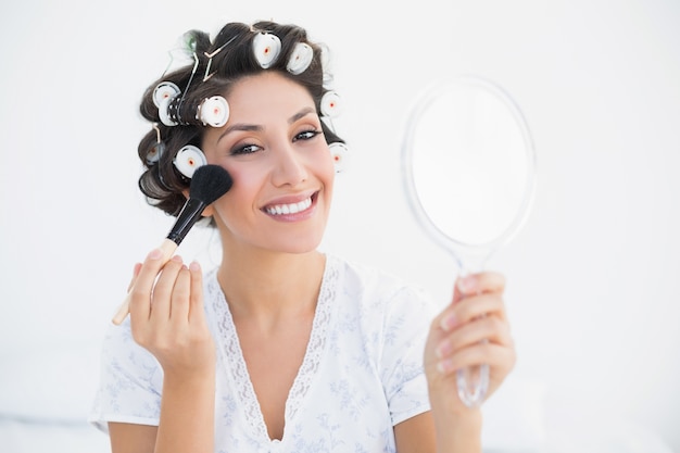 Photo smiling brunette in hair rollers holding hand mirror and applying makeup