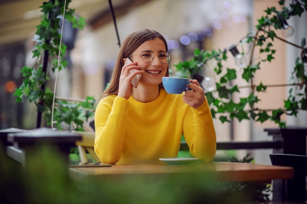 Smiling brunette sitting on terrace of cafe, holding cup of coffee and chatting with friend on a phone.