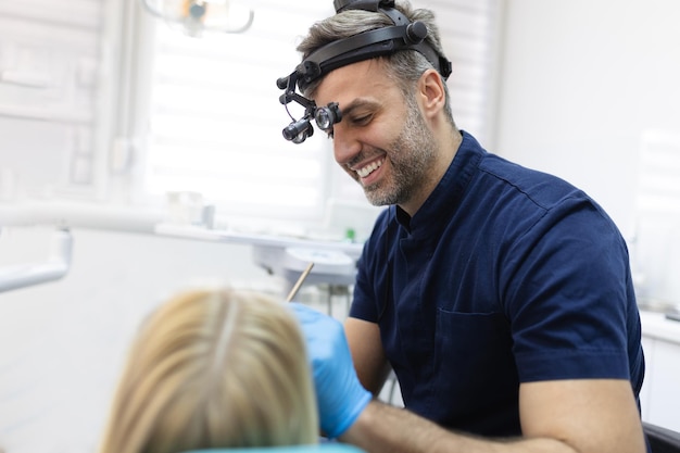 歯科医院で歯科医によって検査されている笑顔のブルネットの女性 患者の口の近くに歯科用器具を保持している医師の手 健康な歯と医学の概念