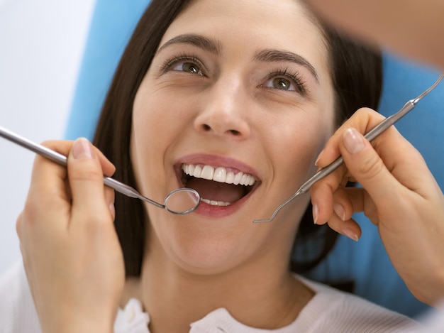 Улыбающаяся брюнетка проходит обследование у дантиста в стоматологической клинике. Руки врача, держащего стоматологические инструменты возле рта пациента. Здоровые зубы и концепция медицины.