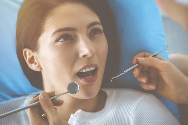 Smiling brunette woman being examined by dentist at dental\
clinic. hands of a doctor holding dental instruments near patient\'s\
mouth. healthy teeth and medicine concept