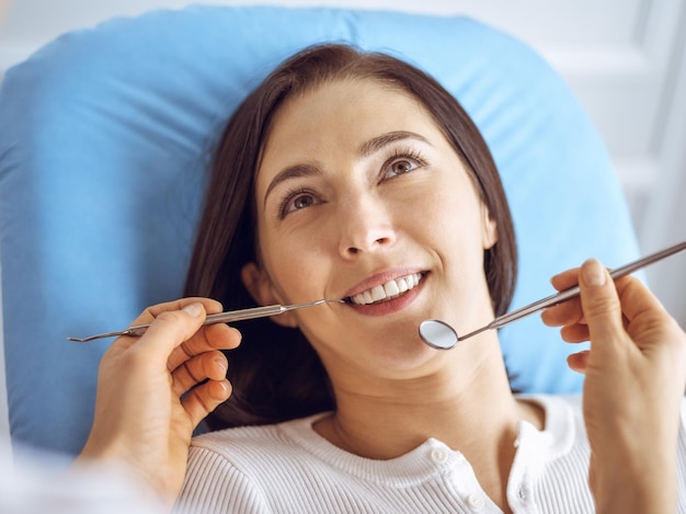 Улыбающаяся брюнетка проходит обследование у дантиста в стоматологической клинике. Руки врача, держащего стоматологические инструменты возле рта пациента. Здоровые зубы и концепция медицины.