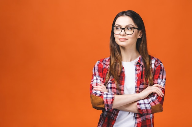 Smiling brunette woman in eyeglasses and casual posing with crossed arms and looking at the camera over orange background.