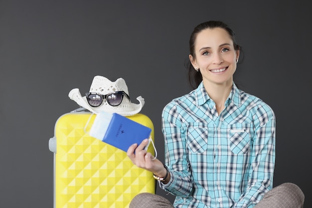 Smiling brunette woman holding passport with ticket and mask\
next to suitcase tourist travel