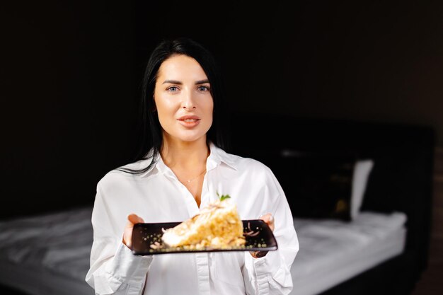 Smiling brunette woman holding plate with some cake in the bedroom in her apartment