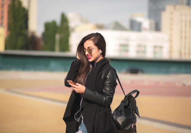 Smiling brunette woman tourist in glasses listen music on cell telephone, standing at the street