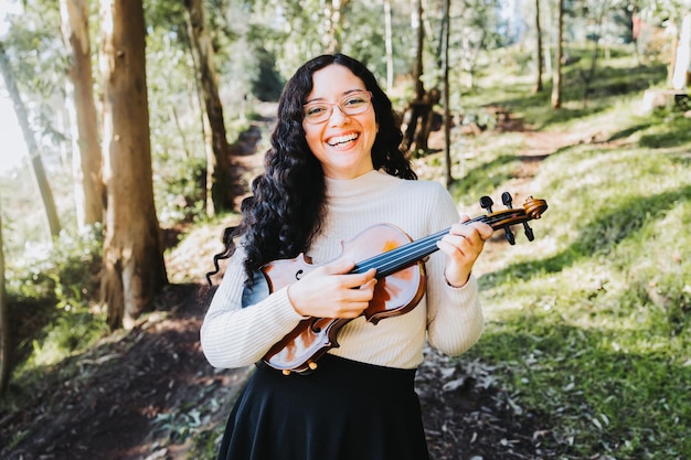 Donna bruna sorridente con gli occhiali in possesso di un violino fuori nel bosco