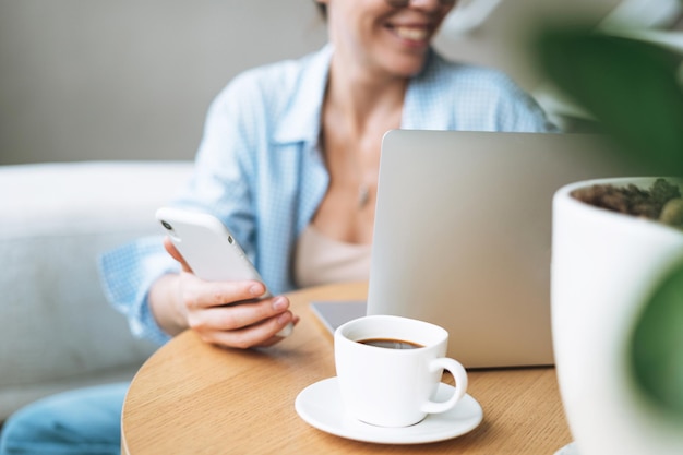 Smiling brunette woman with long hair working on laptop using mobile phone in bright home