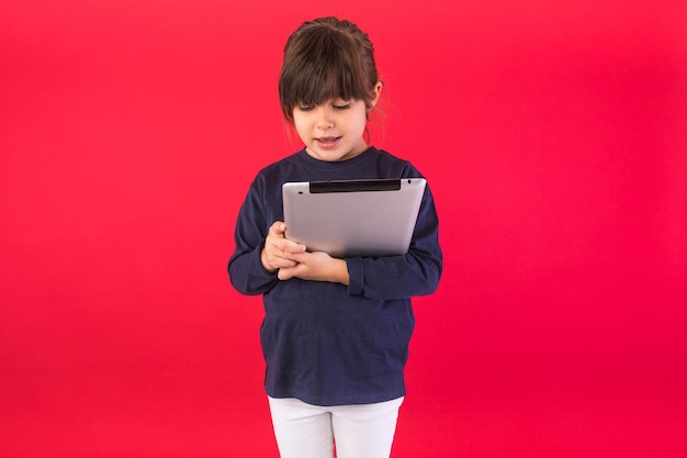 Smiling brunettehaired girl wearing blue sweatshirt and white pants holding and looking at digital tablet on red background Concept of call center call speak and listen