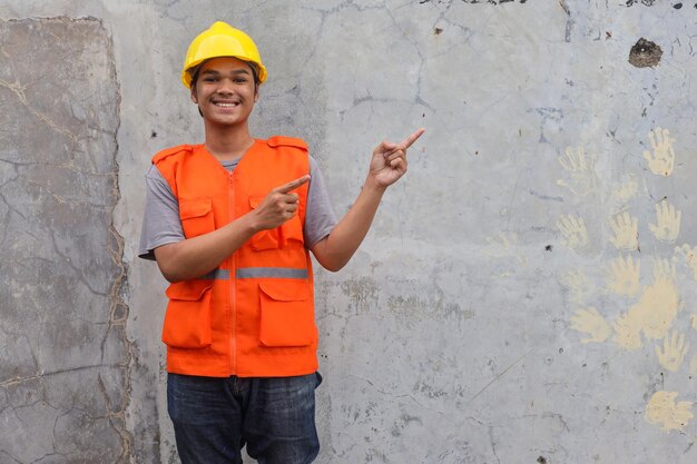 Smiling builder construction guy in yellow helmet and vest pointing fingers aside at free space stan