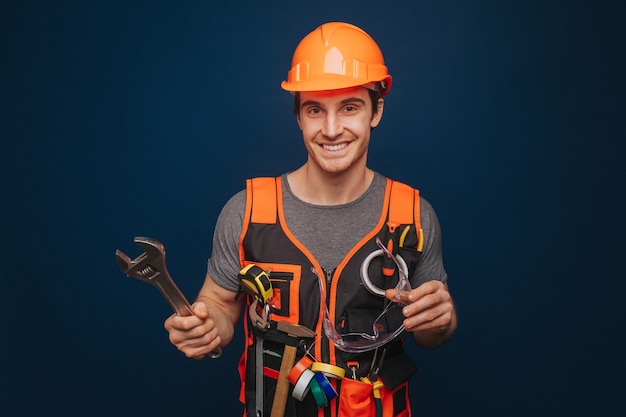 Smiling builder in a helmet is holding glasses and wrench