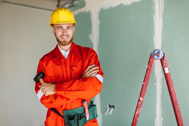 Smiling builder in orange work clothes and yellow hardhat holding hammer in hand  joyfully looking in camera with ladder on background