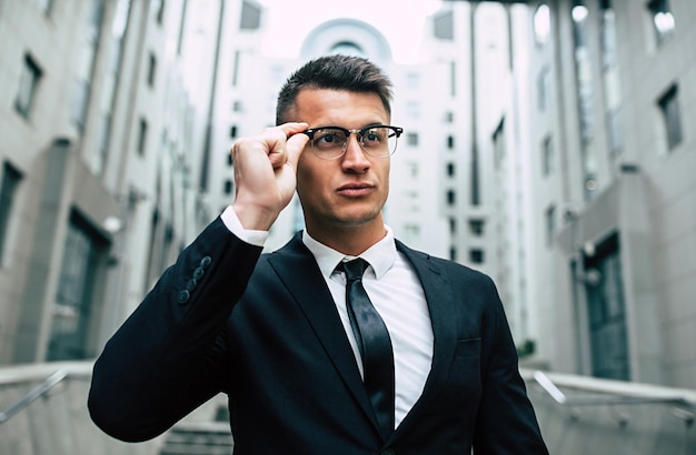 Smiling business man in full black suit and glasses looking on camera standing on cityscape background