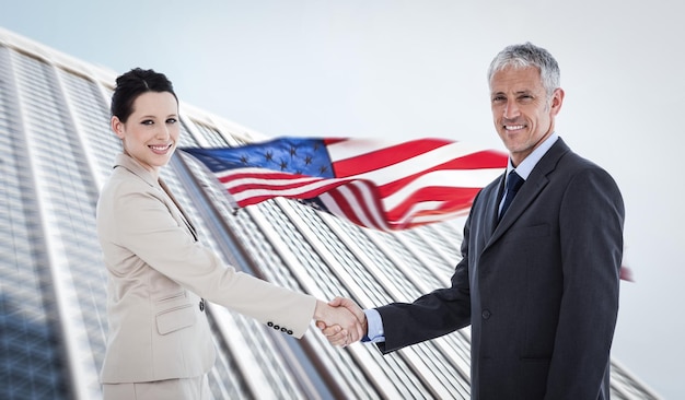 Smiling business people shaking hands while looking at the camera against american flag and skyscraper