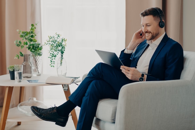 Smiling business person in suit wearing headset having video call, working online on laptop while sitting in living room in comfortable armchair. Business people and remote work at home