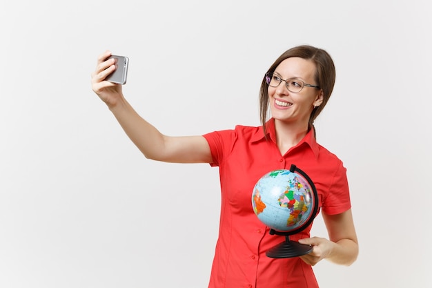 Smiling business teacher woman in red shirt holding mobile phone and doing taking selfie shot with globe isolated on white background. Education teaching in high school university concept. Copy space.