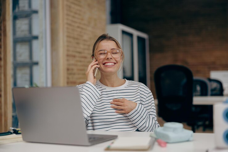 Photo smiling business woman in eyeglasses working on laptop while sitting in office and talking by phone