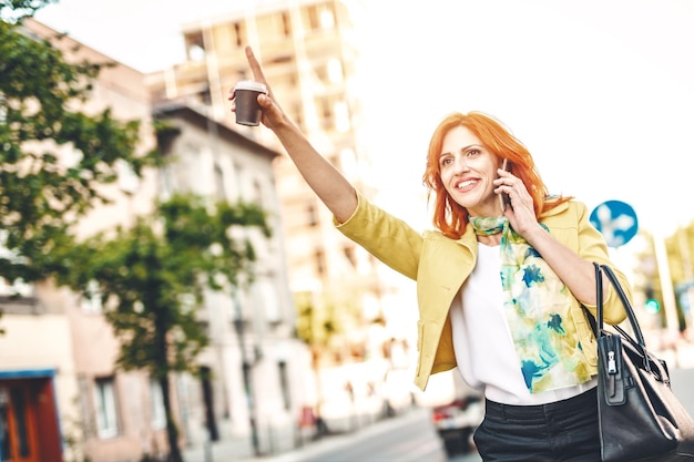 Photo smiling business woman is standing on the street with smartphone and calling for taxi.