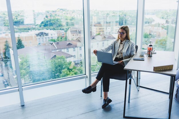 Улыбающаяся деловая женщина смотрит на ноутбук во время работы в офисе Концепция современной успешной женщины Идея бизнеса и жизни предпринимателя Молодая женщина за столом с открытым ноутбуком
