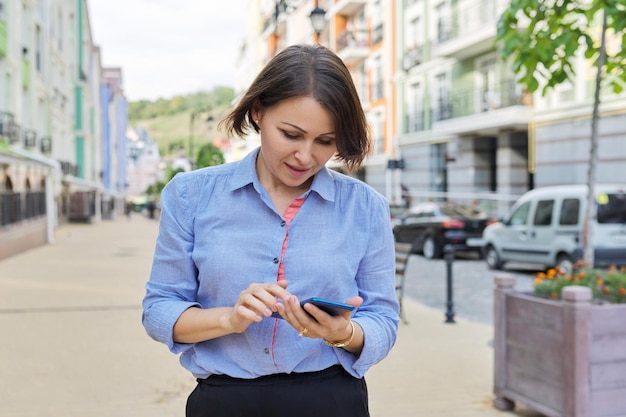 Donna d'affari sorridente che legge la digitazione guardando lo schermo dello smartphone camminando lungo la strada della città