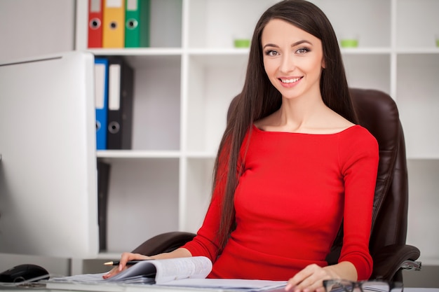 Smiling business woman in red dress sitting and writing something by the table as well as looking at camera