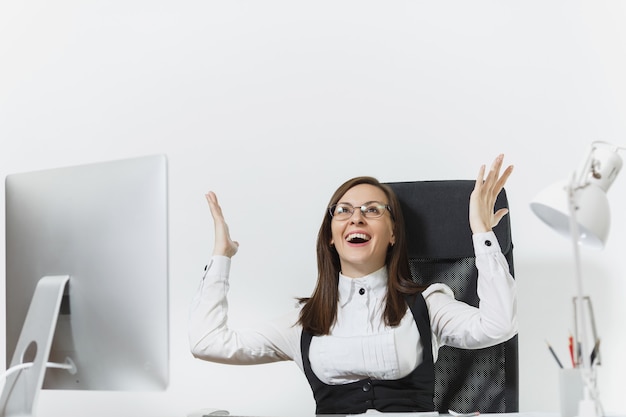 Smiling business woman sitting at the desk, working at computer with modern monitor and documents in office, rejoicing at success, holding hands up, copy space for advertisement