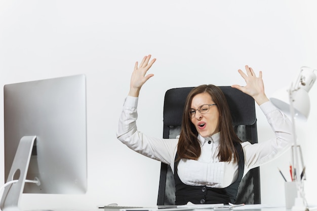 Smiling business woman sitting at the desk, working at computer with modern monitor and documents in office, rejoicing at success, holding hands up, copy space for advertisement