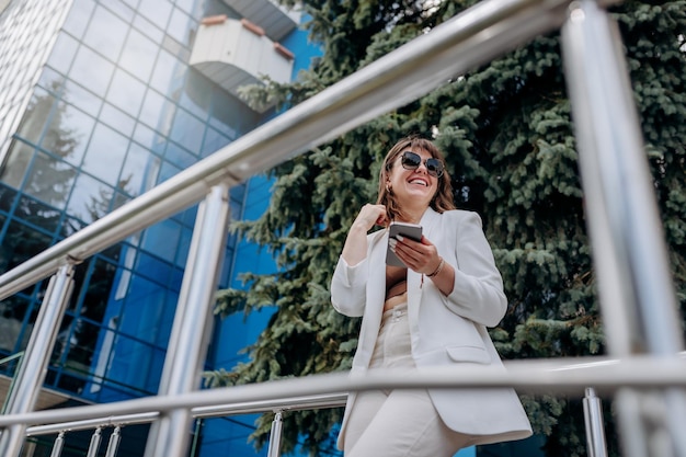 Smiling business woman in white suit and sunglasses using phone during break standing near modern office building