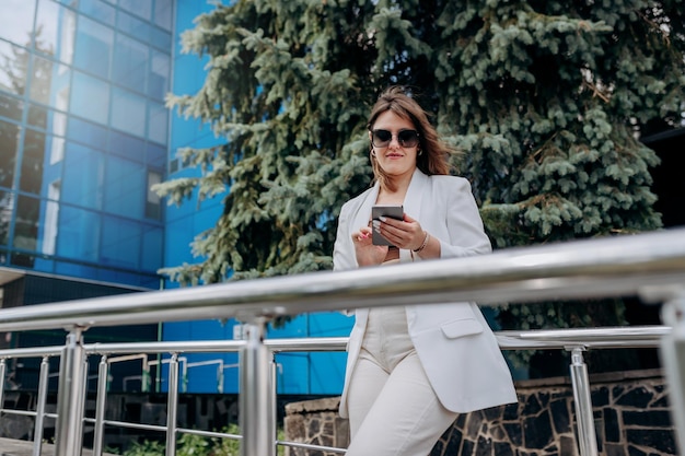 Donna d'affari sorridente in abito bianco e occhiali da sole che usa il telefono durante la pausa in piedi vicino a un moderno edificio per uffici