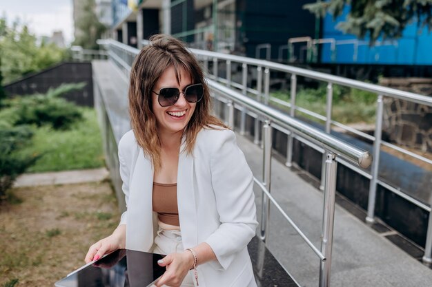 Smiling business woman in white suit and sunglasses working on digital tablet sitting near modern office building