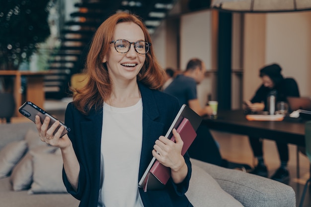 Smiling business woman with modern smartphone in hand, sitting on couch in lounge zone, holding laptop with notebook, smiling and looking around while waiting for meeting with client in office space