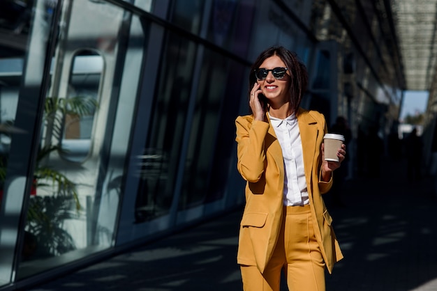 Smiling business woman in yellow suit walking near business centre at the city, communicates via smartphone and drinks coffee. Business concept, communication, banker, manager and office worker.